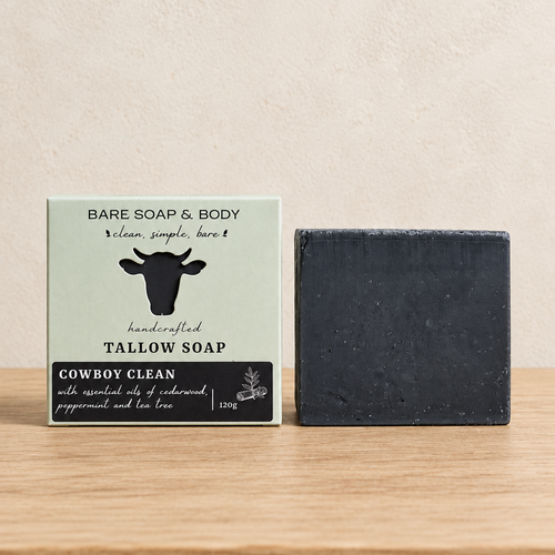 A bar of Bare Soap & Body tallow soap sits next to its packaging box on a light wooden surface against a textured beige wall. The soap bar is a dark charcoal gray, and the box is a pale sage green with a black cow logo and black text.