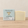 A bar of white handmade soap stands next to its light blue and white packaging box on a light wood surface against a textured beige wall. The box is labeled 'Bare Soap & Body' and 'Magnesium'.