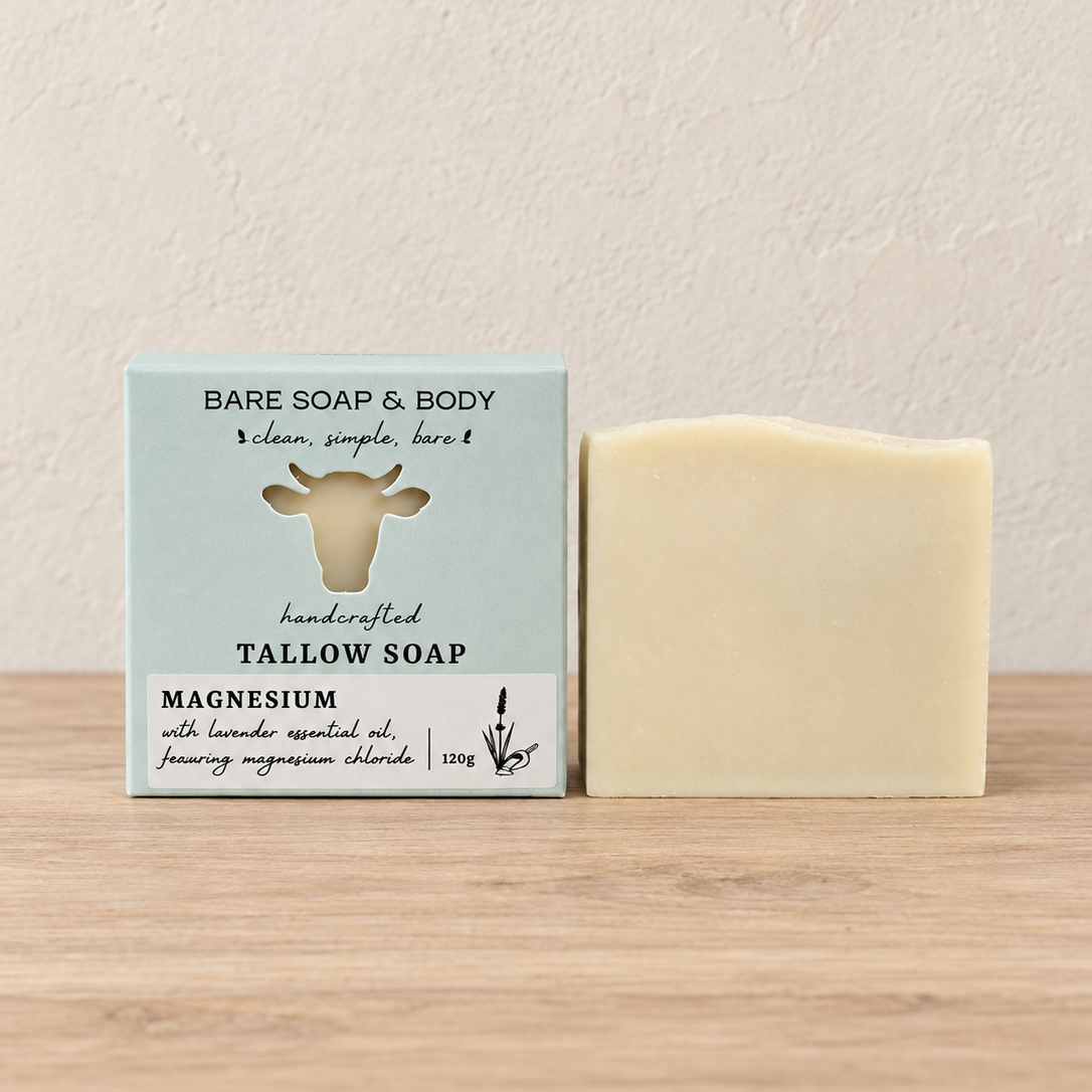 A bar of white handmade soap stands next to its light blue and white packaging box on a light wood surface against a textured beige wall. The box is labeled 'Bare Soap & Body' and 'Magnesium'.