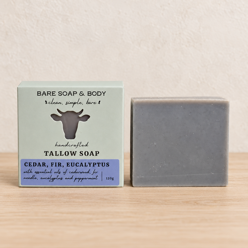 A bar of Bare Soap & Body Tallow Soap stands next to its packaging box on a light wooden surface against a textured beige wall. The soap is a muted gray, and the box is pale sage green with a purple label and a black cow graphic.