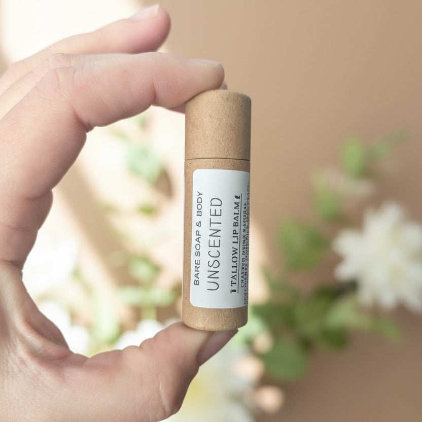A hand holding a small cylindrical tube labeled 'UNSCENTED' in front of a blurred indoor room background.