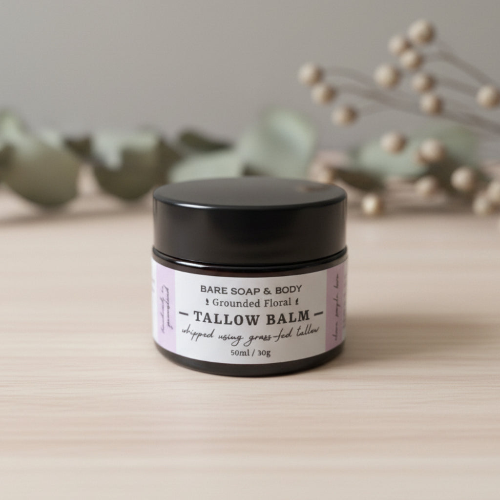 A small black jar of Tallow Balm by Bare Soap & Body, featuring a white label with black text, set on a gray tabletop with a blurred background.