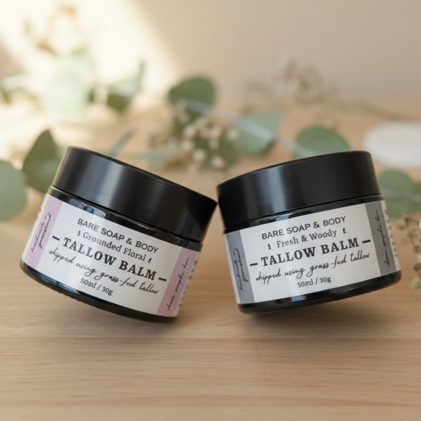 Two small black jars of Bare Soap & Body Tallow Balm, one labeled Fresh & Woods 1 and the other Grounded Finish 1, placed on a metallic surface with the lid off in the background.