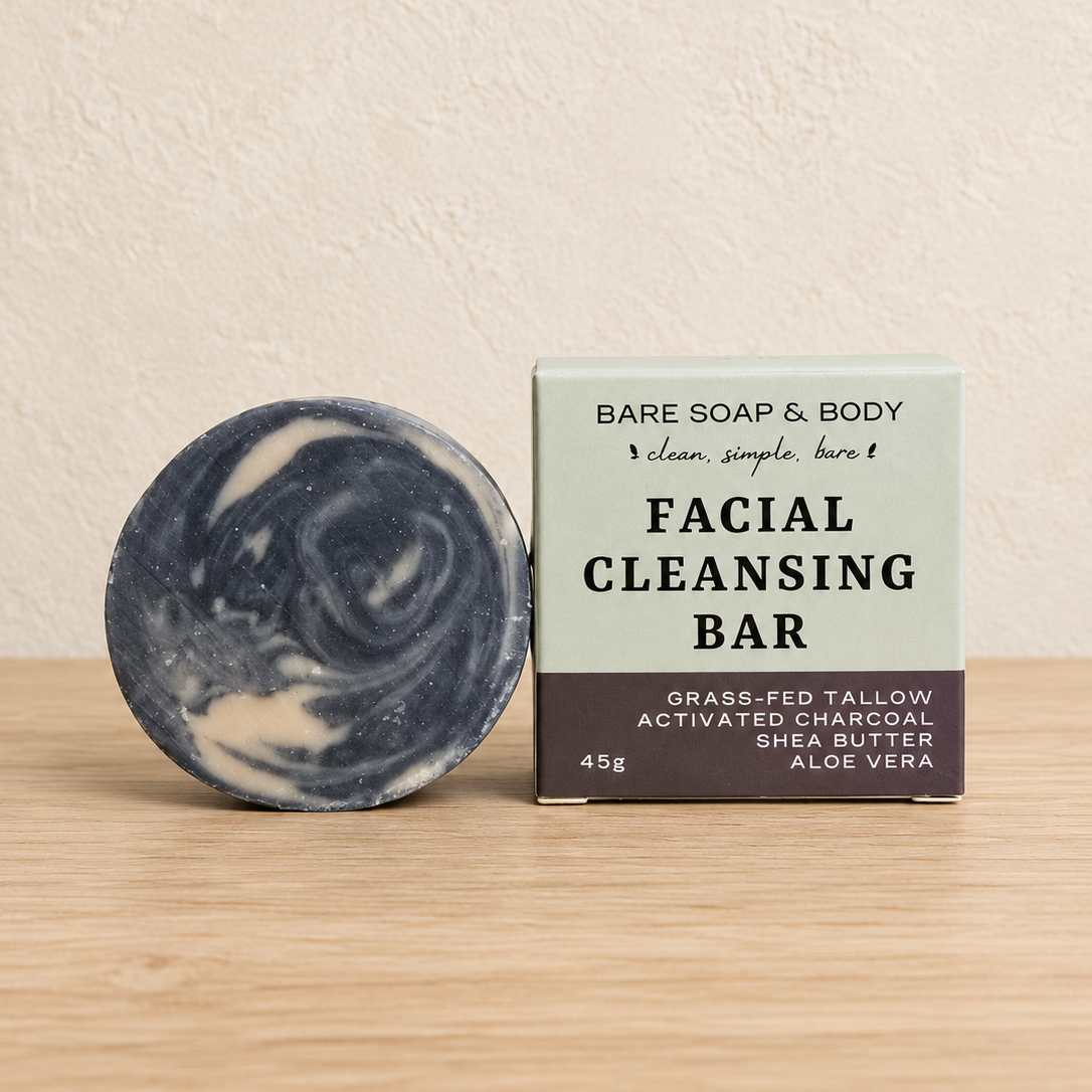 A round marbled facial cleansing bar soap next to its packaging box on a light wood surface against a textured beige wall. The box is labeled 'BARE SOAP & BODY FACIAL CLEANSING BAR'.