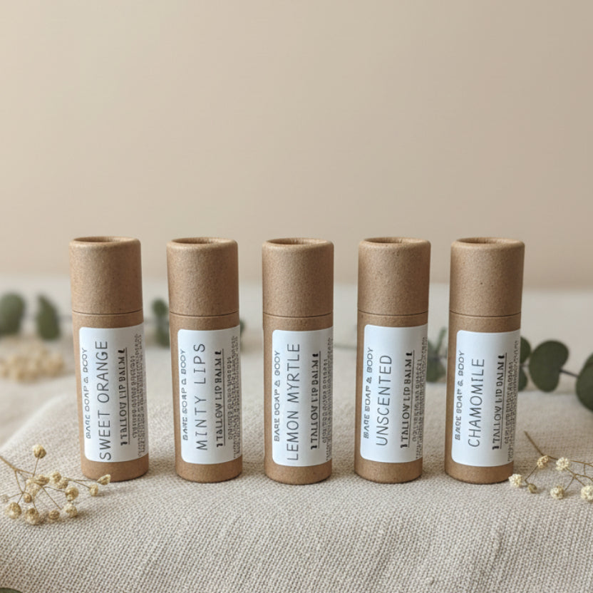 Six lip balm tubes standing upright on a wooden surface against a light wall. The tubes are brown kraft paper with white labels showing scented lip balm varieties including Sweet Orange, Minty Lips, Lemon Myrtle, Unscented, and Chamomile.