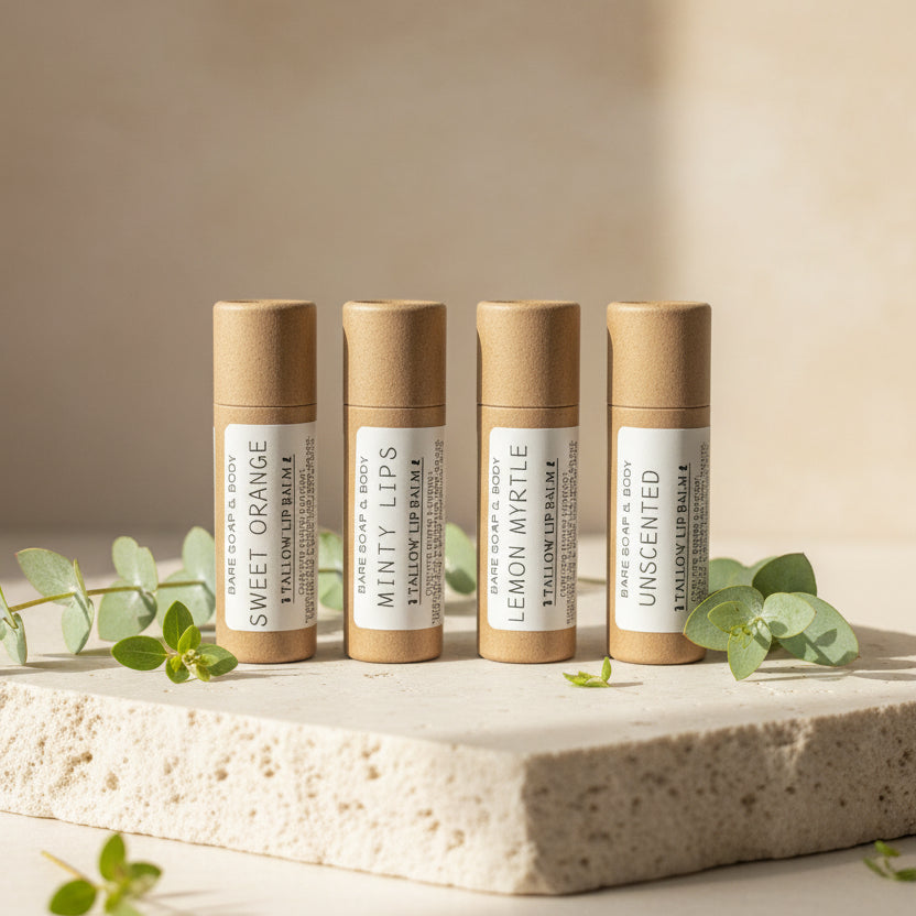 Four small lip balm tubes standing upright on a wooden surface against a plain light background. The labels read Sweet Orange, Minty Sips, Lemon Myrtle, and Unscented.