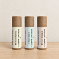 Three bare lip balm tubes standing upright on a light wood surface against a textured off-white wall. The tubes have kraft brown caps and cream labels with black text, labeled Lemon Myrtle, Minty Lips, and Sweet Orange.