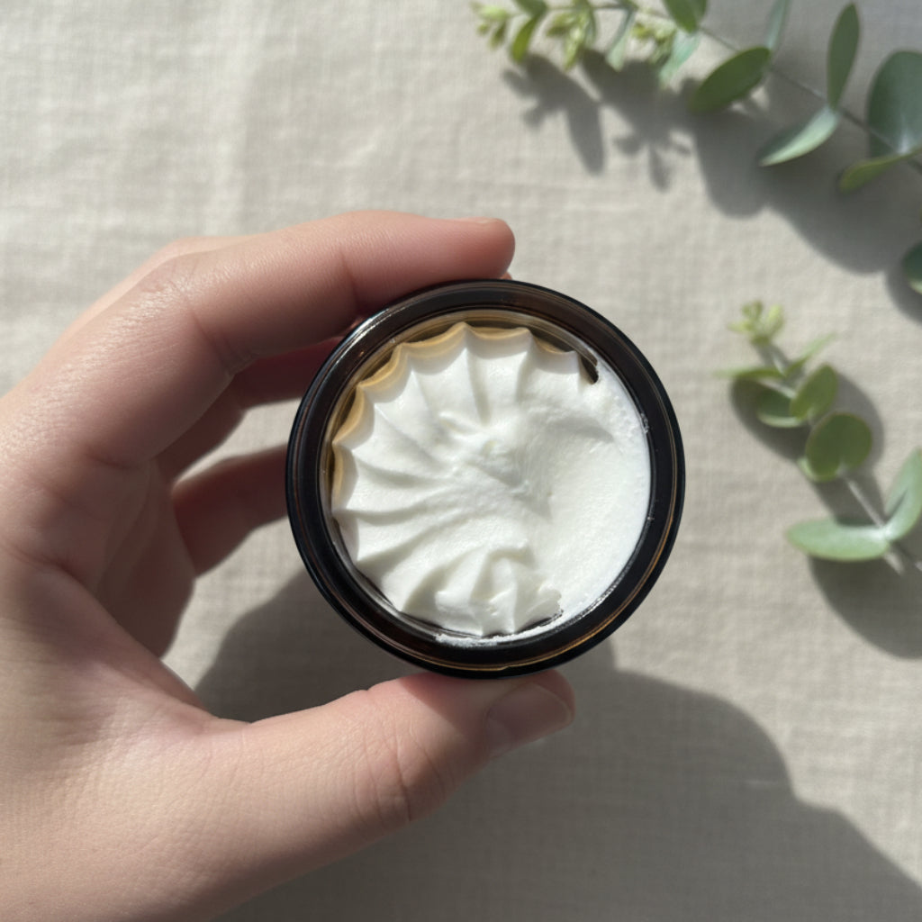 A hand holding a small round cosmetic container filled with white cream or powder, photographed against a plain indoor background.