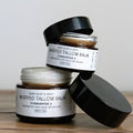 Two jars of Whipped Tallow Balm in amber jars with black lids on a wooden surface.