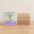 A handmade tallow soap bar in a two-tone beige and light brown color sits next to its packaging box on a light wooden surface against a textured cream wall. The box is pale green with a cow illustration and purple accents, labeled 'Bare Soap & Body' and 'In the Garden'.