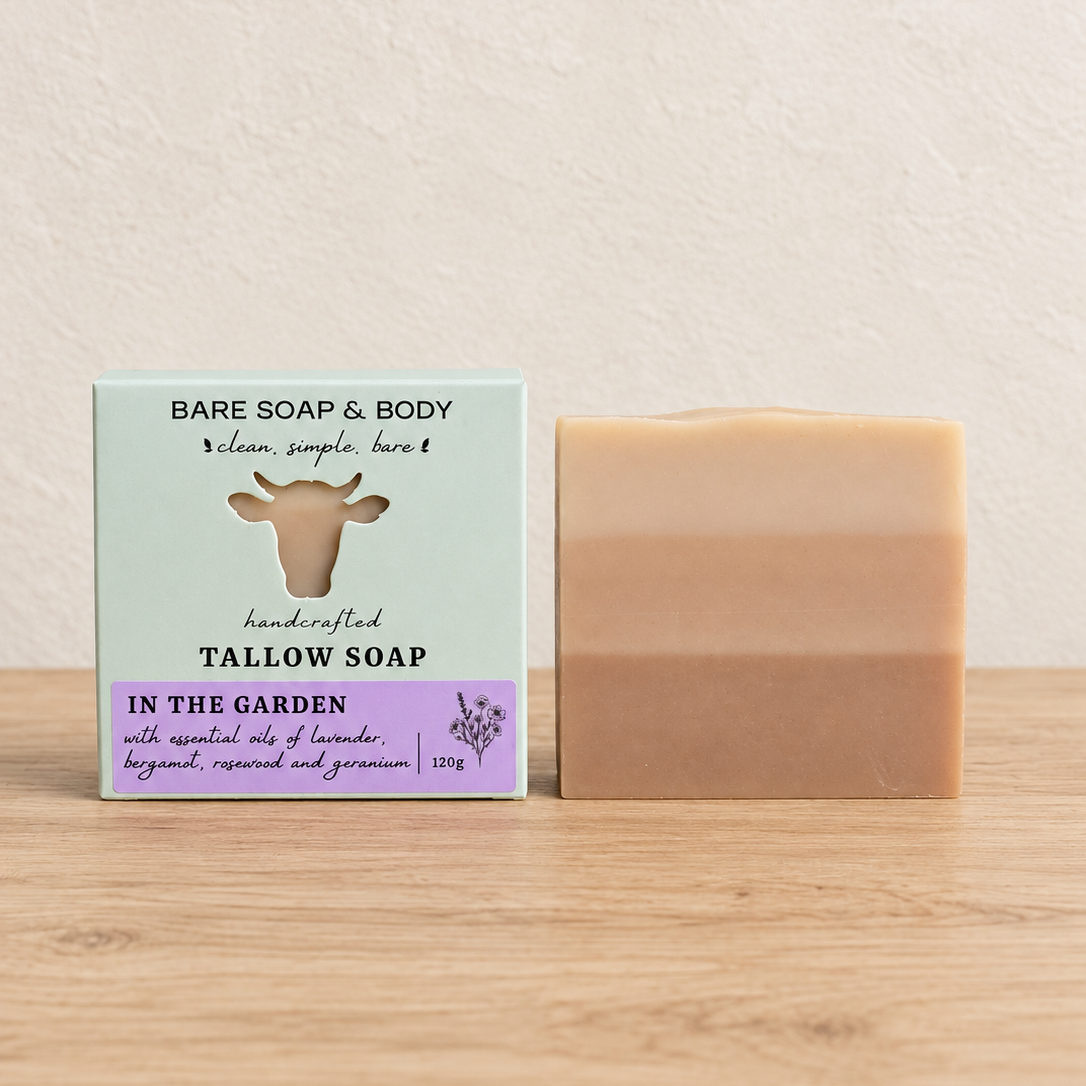 A handmade tallow soap bar in a two-tone beige and light brown color sits next to its packaging box on a light wooden surface against a textured cream wall. The box is pale green with a cow illustration and purple accents, labeled 'Bare Soap & Body' and 'In the Garden'.