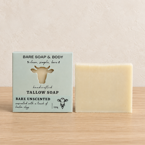 A bar of pale yellow tallow soap stands next to its light blue-green packaging box on a light wood surface against a textured beige wall. The box features a cow silhouette and the text 'BARE SOAP & BODY' and 'BARE UNSCENTED'.