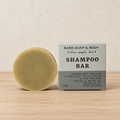 A round bar of shampoo soap next to its packaging box on a light wood surface against a textured beige wall. The box is labeled 'BARE SOAP & BODY SHAMPOO BAR'.