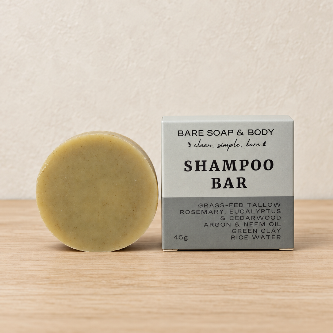 A round bar of shampoo soap next to its packaging box on a light wood surface against a textured beige wall. The box is labeled 'BARE SOAP & BODY SHAMPOO BAR'.