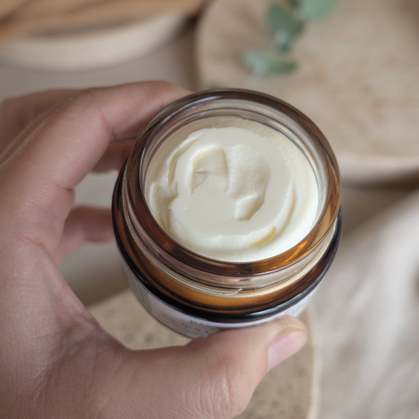 A hand holding an open amber glass jar filled with a beautiful whipped tallow balm, photographed in a real indoor setting with a speckled countertop and blurred background.
