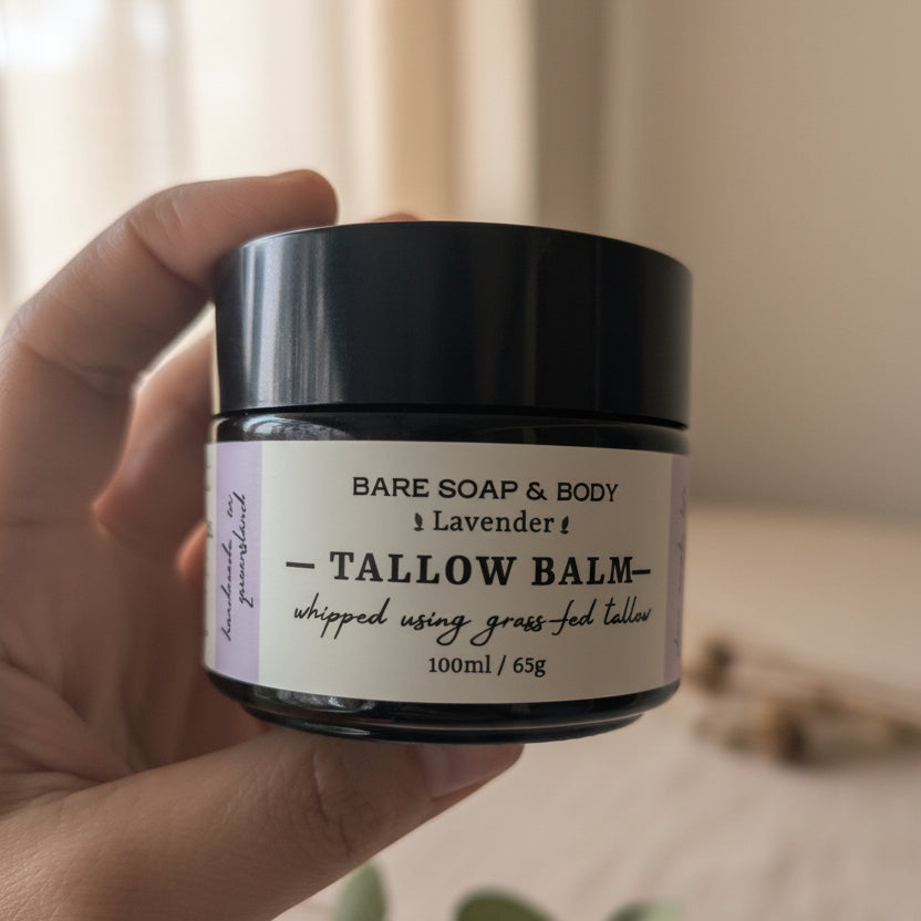 A hand holding a jar of Bare Soap & Body Tallow Balm in lavender, featuring a black lid and a cream-colored label with black text, set against a blurred outdoor background.