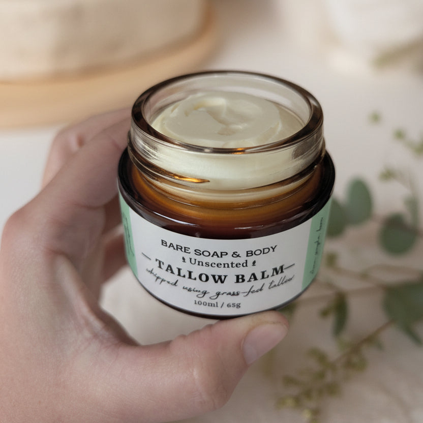 A hand holding an open jar of Bare Soap & Body Tallow Balm, showing the creamy off-white balm inside a clear amber glass jar with a white and green label.