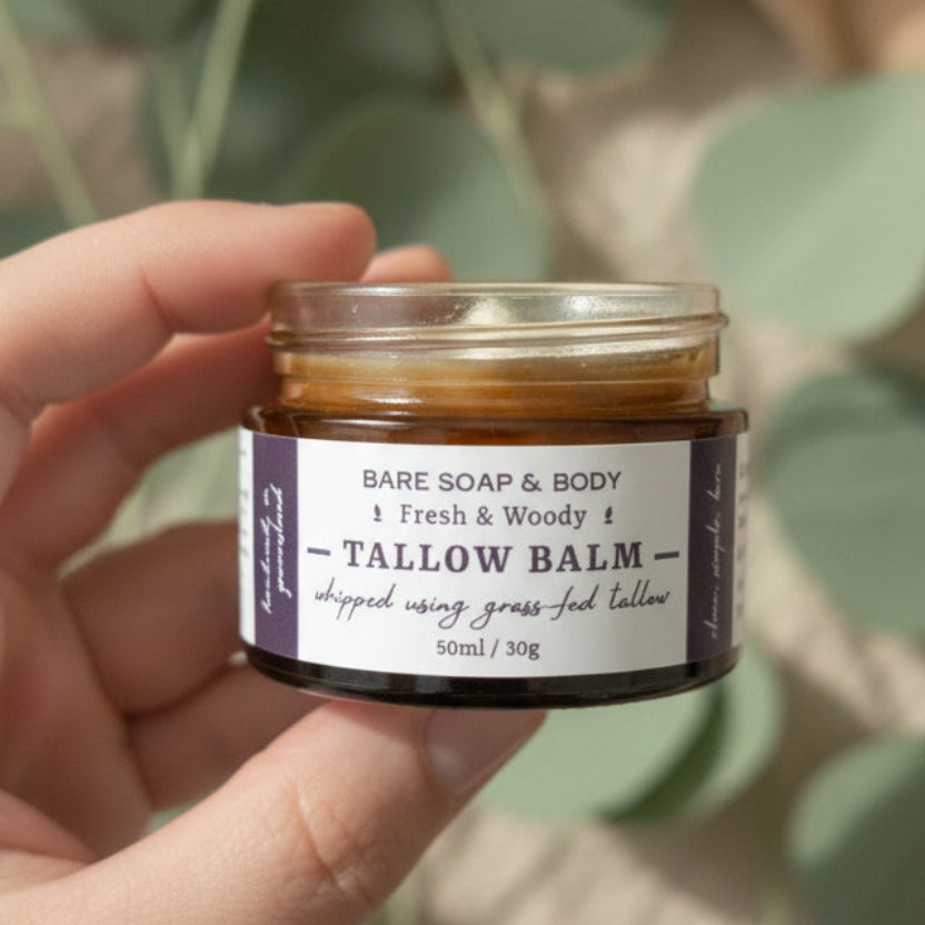 A hand holding a small glass jar of Tallow Balm by Bare Soap & Body. The jar has a clear lid and a white label with black text, showing the 'b' logo on the side.