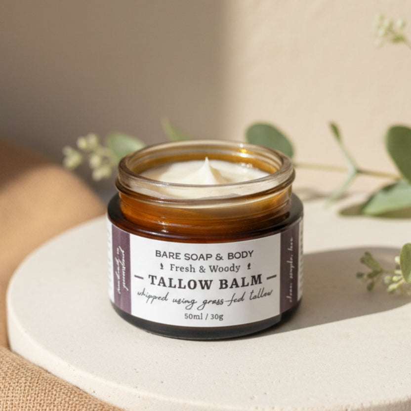 A hand holding a small glass jar of Bare Soap & Body Tallow Balm with a white label, showing the white balm inside against a plain indoor background.