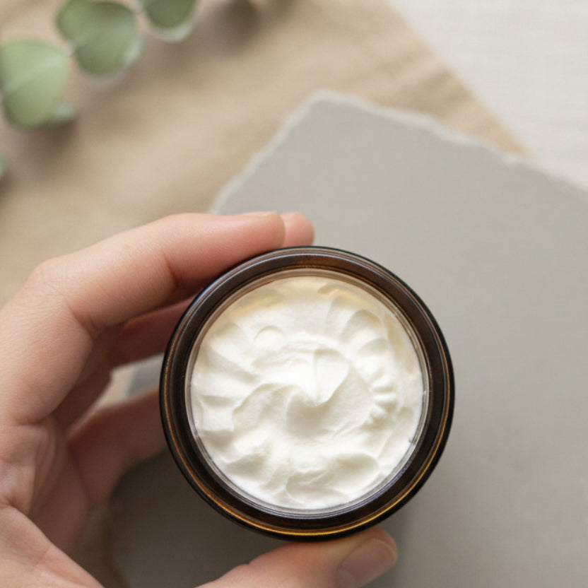 A hand holding a small round glass jar filled with a thick white cream or balm, photographed against a plain indoor background.